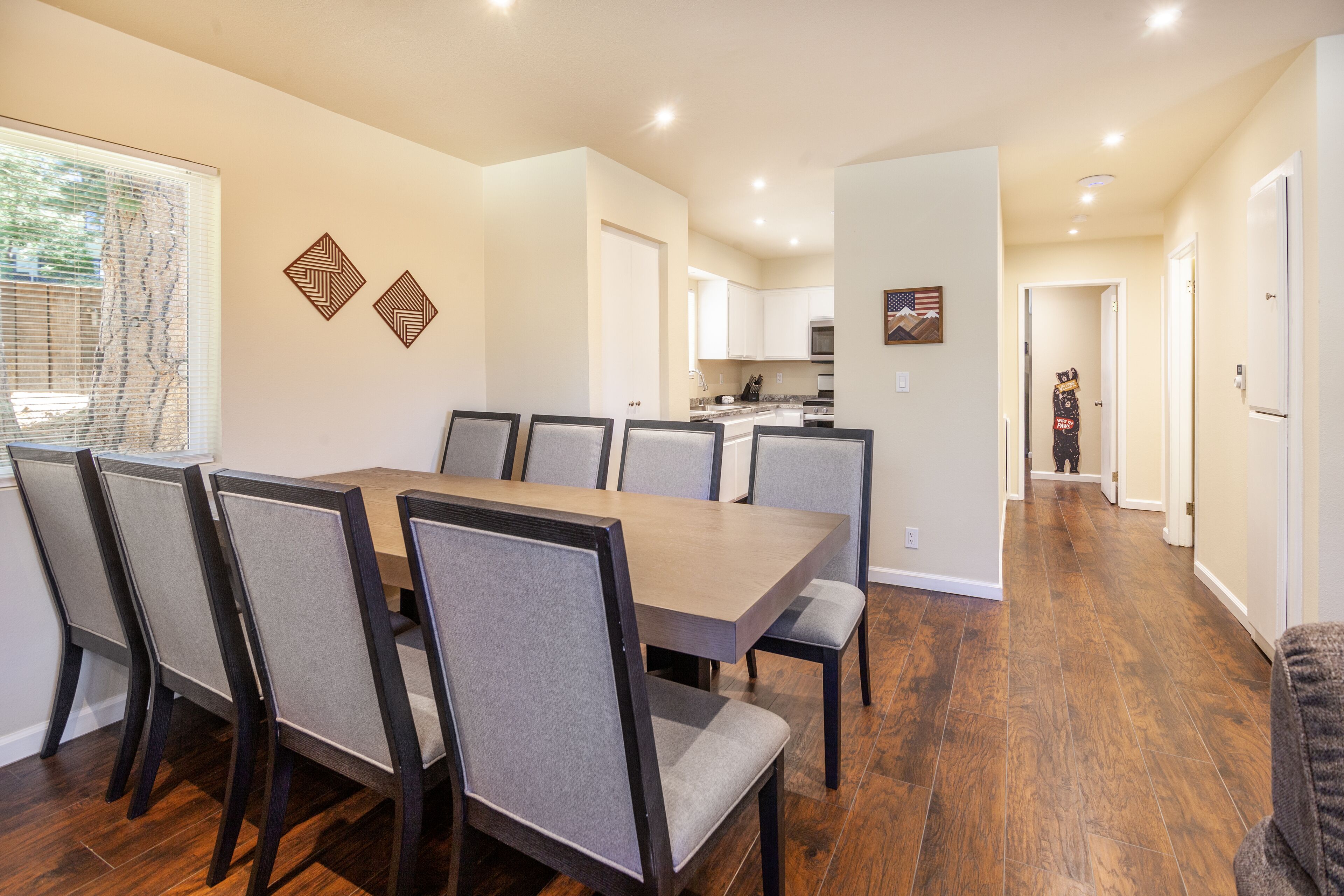 Un coin repas avec une table en bois et six chaises, une cuisine à l’arrière-plan et un couloir menant à une autre pièce.