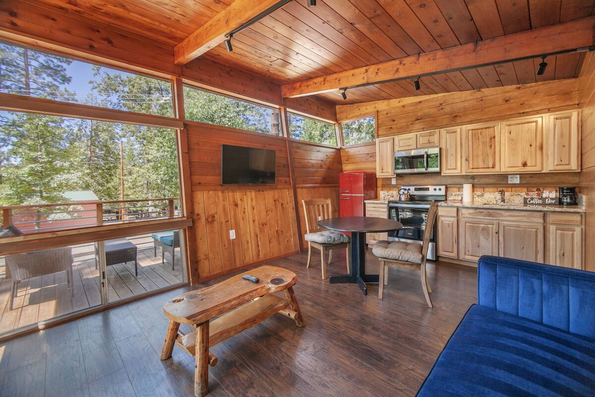 A wooden cabin interior with a dining area, kitchen, and a large window overlooking the forest.