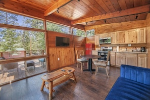 A wooden cabin interior with a dining area, kitchen, and a large window overlooking the forest.