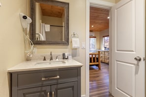 A bathroom with a sink, mirror, and hairdryer. There is a towel rack with a towel hanging on it. The bathroom has wooden flooring and a door leading to another room.