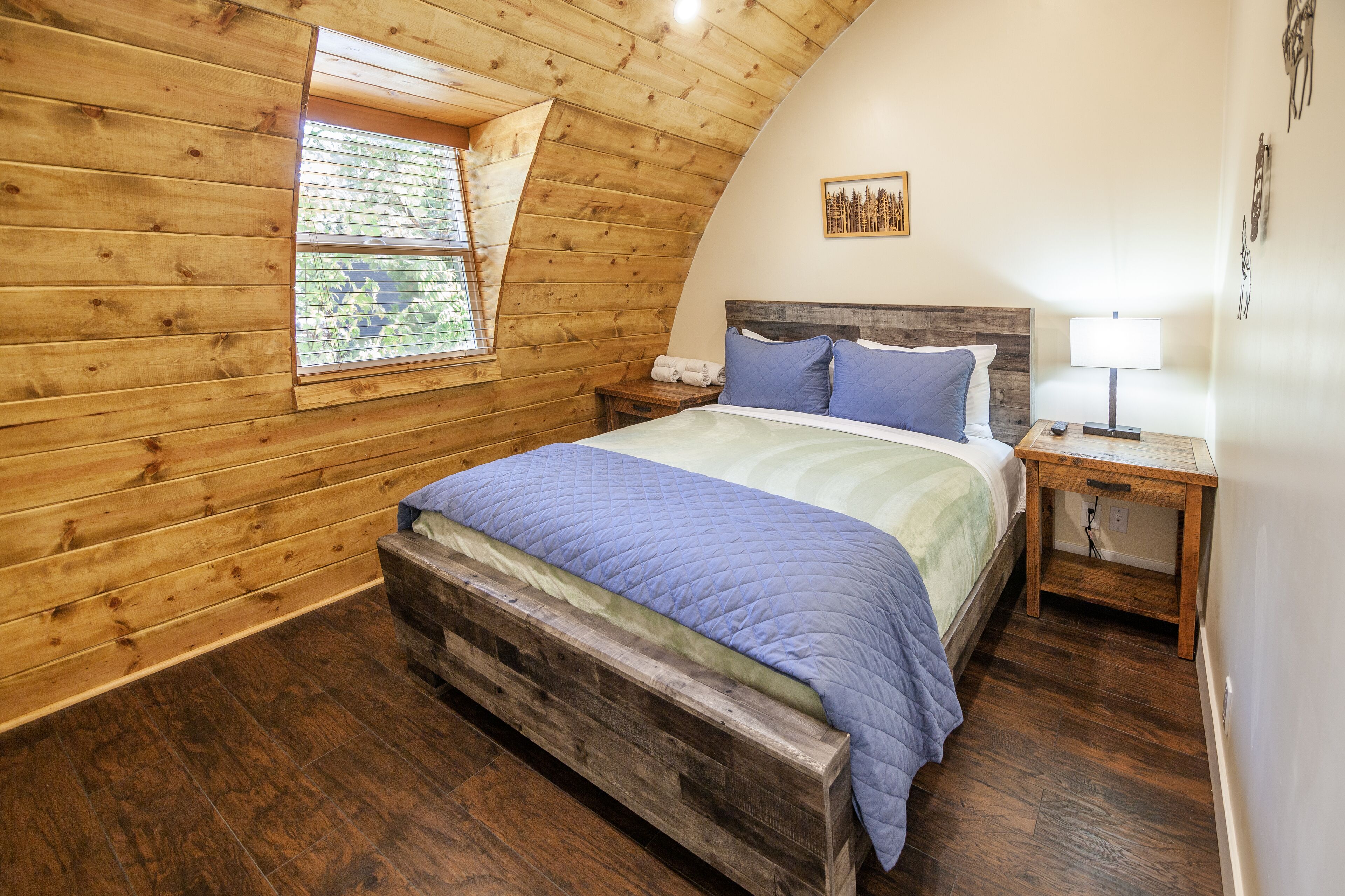 A bedroom with a wooden sloped ceiling, a bed with a blue quilt, a nightstand with a lamp, and a window with a view of trees.