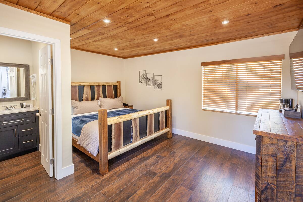 A bedroom with a wooden ceiling, a bed with a wooden frame, a dresser, and a window with blinds.