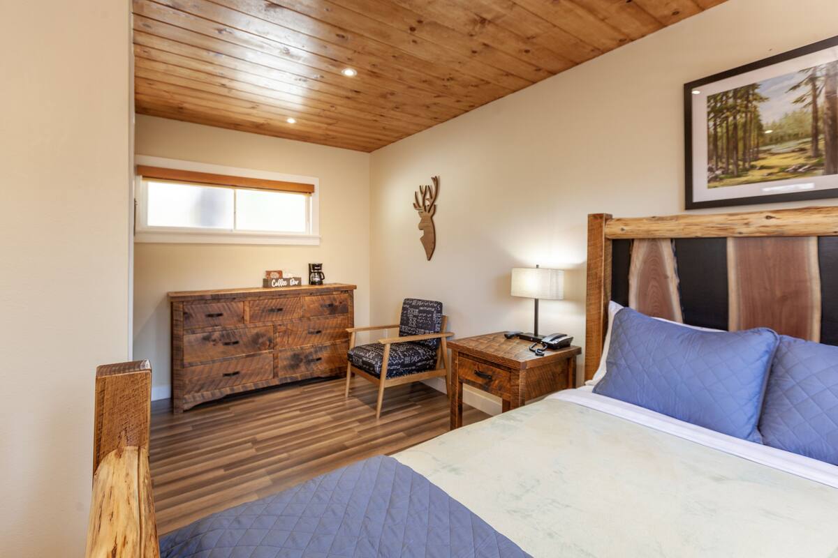 A bedroom with a wooden ceiling, a bed with blue bedding, a wooden dresser, a chair, a desk with a lamp, and a deer head decoration on the wall.