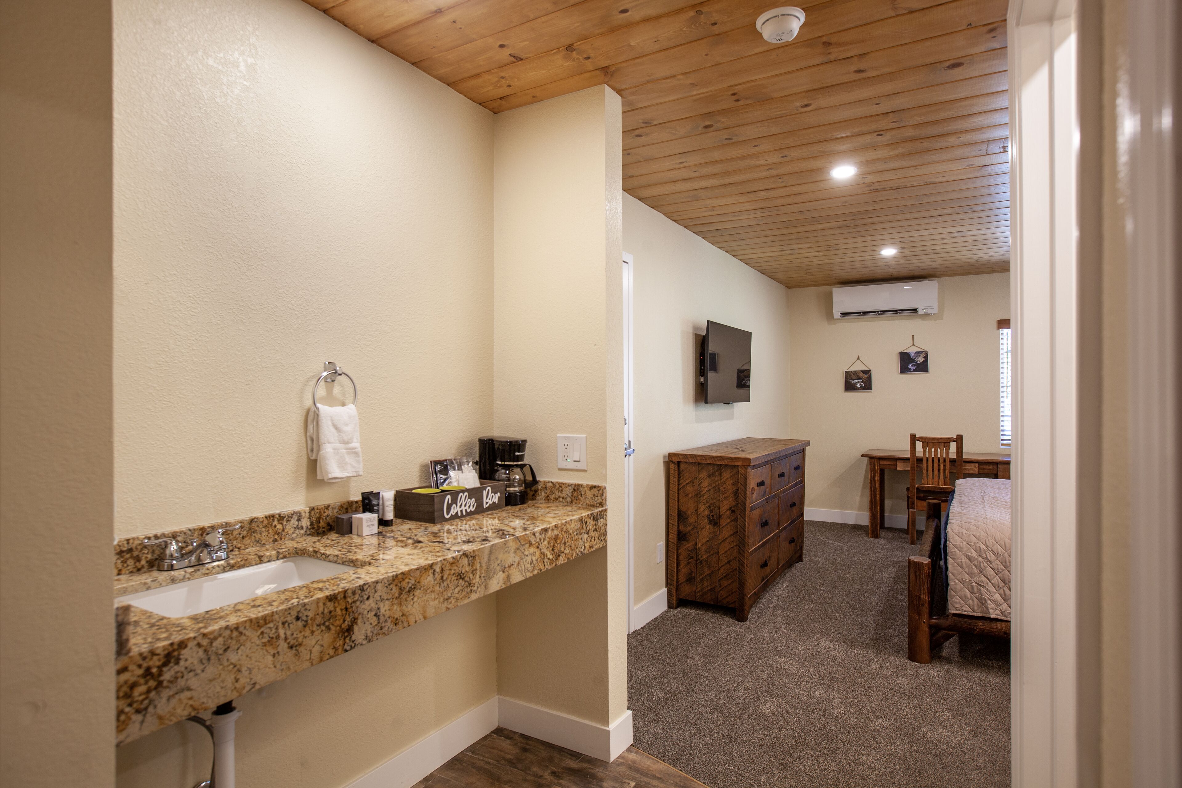 A bathroom with a granite countertop sink, a coffee maker, and a towel rack. There is a wooden dresser and a bed in the adjacent room.