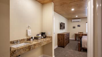 A bathroom with a granite countertop sink, a coffee maker, and a towel rack. There is a wooden dresser and a bed in the adjacent room.