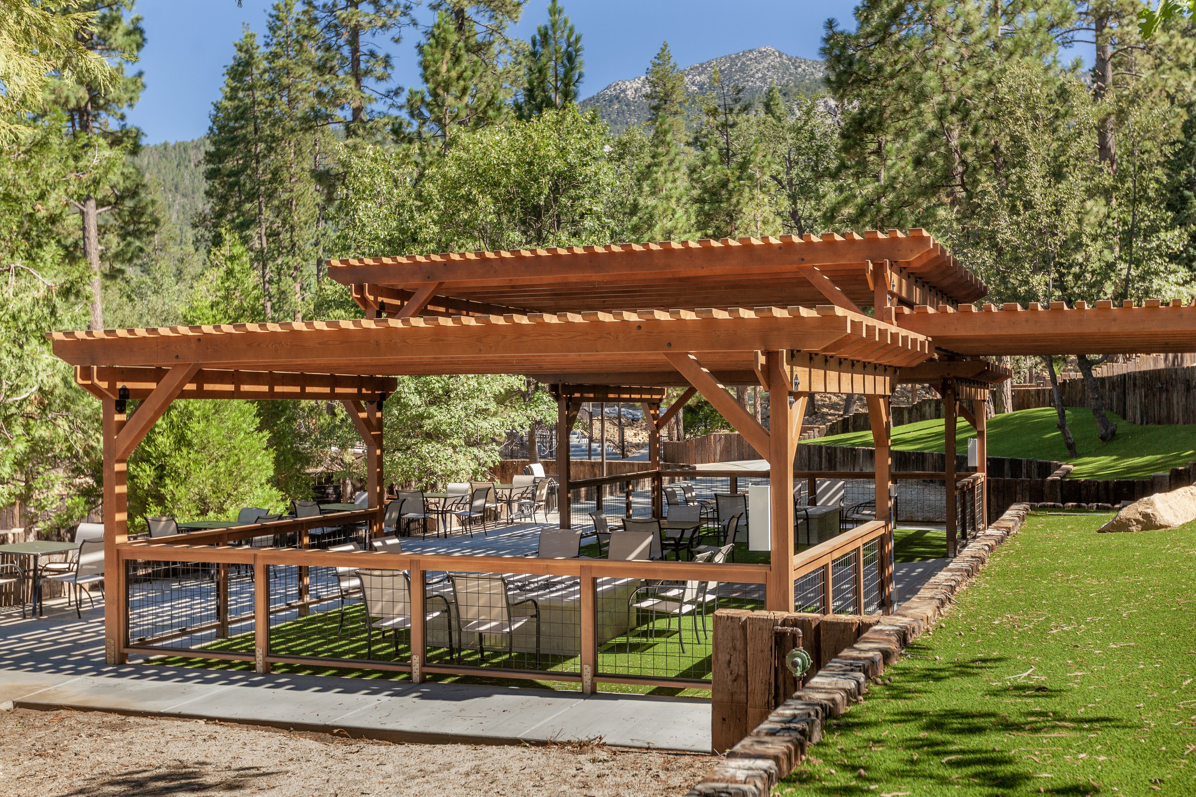 Eine Pergola aus Holz mit Sitzgelegenheiten im Freien, umgeben von einem gepflegten Rasen und dichten Bäumen.