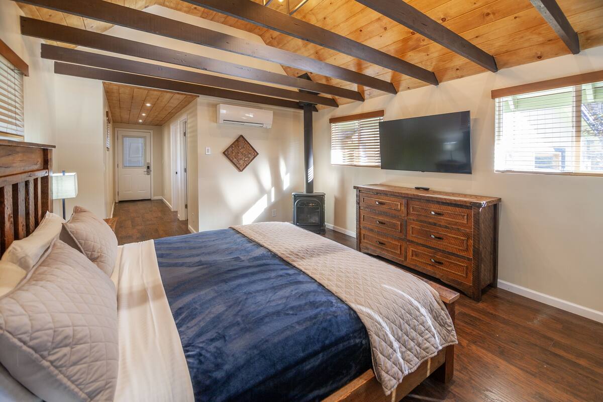 A bedroom with a bed, wooden ceiling beams, a dresser, a TV, and a window with blinds.
