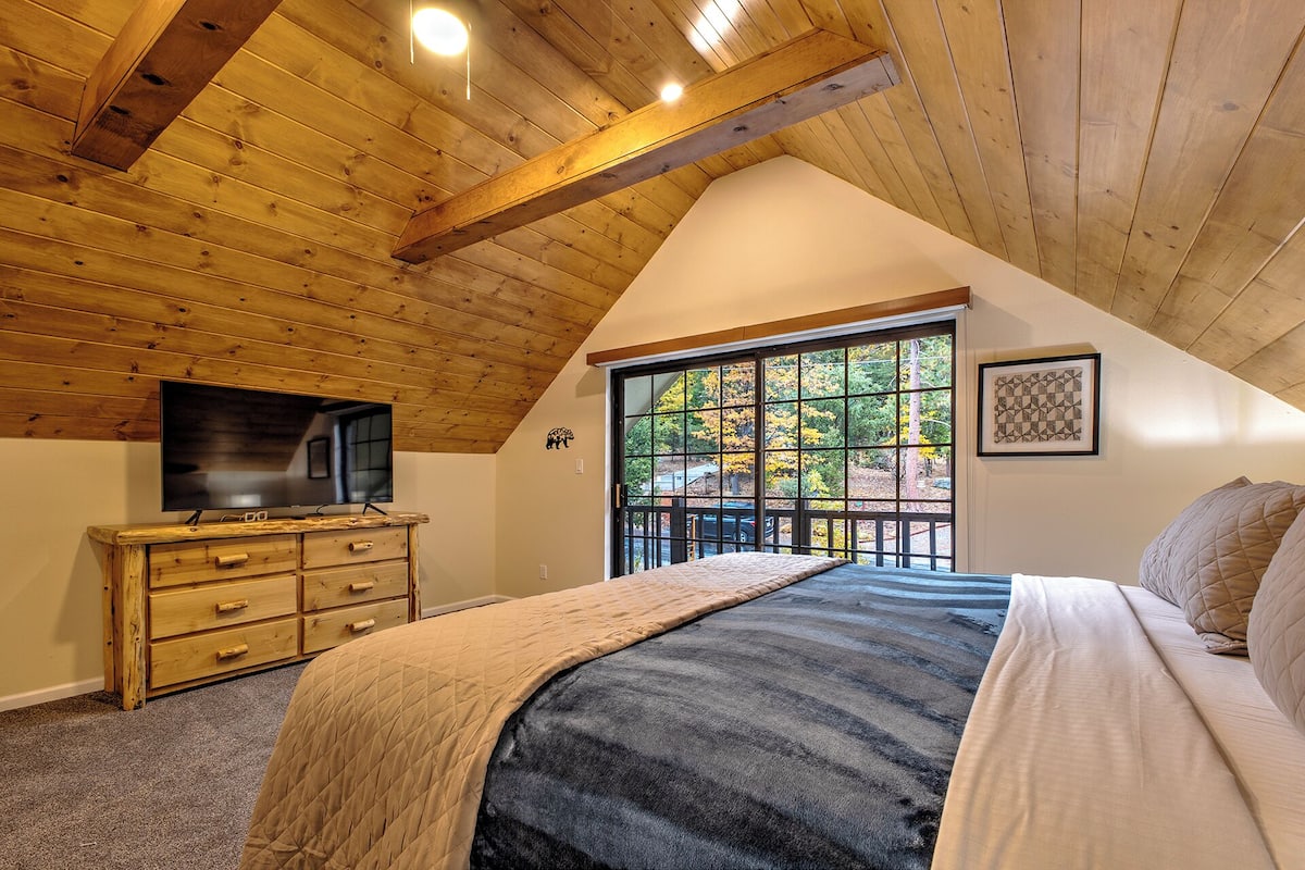 A bedroom with a sloped ceiling, a large window, a bed, a dresser, and a flat-screen TV.
