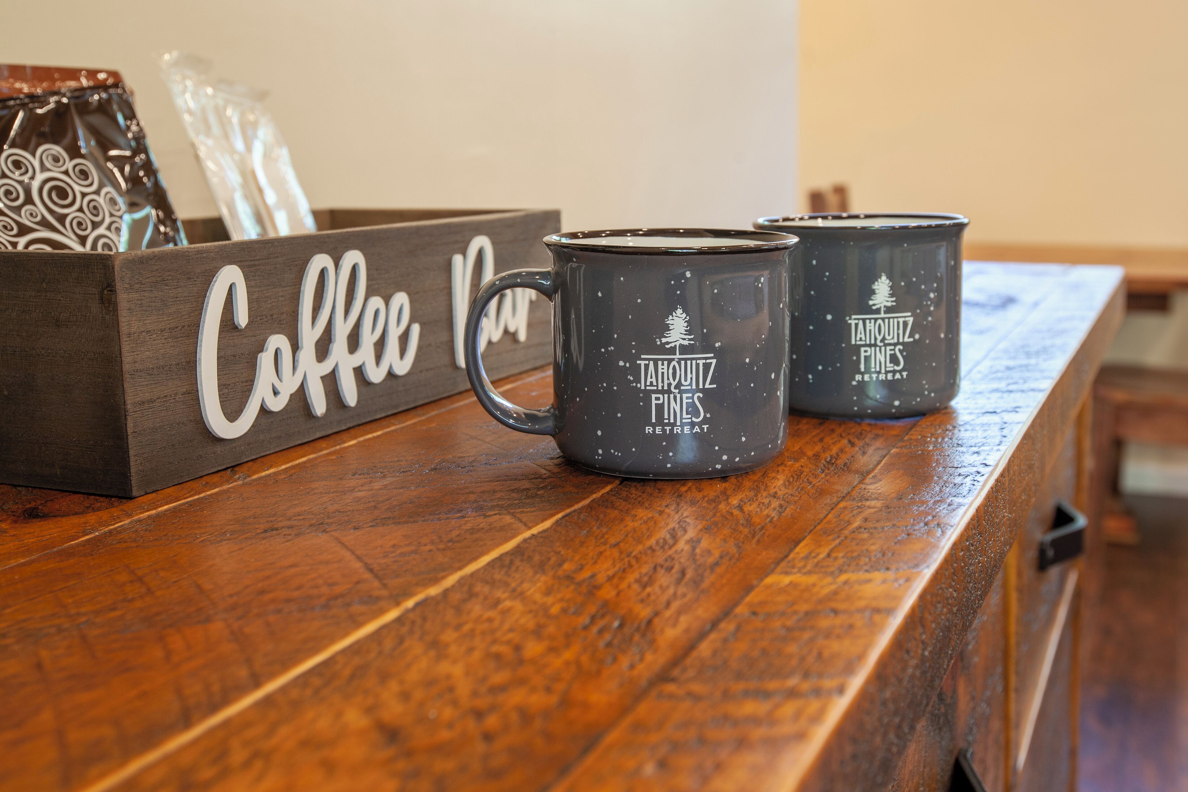 A wooden coffee table with two black mugs labeled "Tahquitz Pines Retreat" and a wooden sign that reads "Coffee Bar".