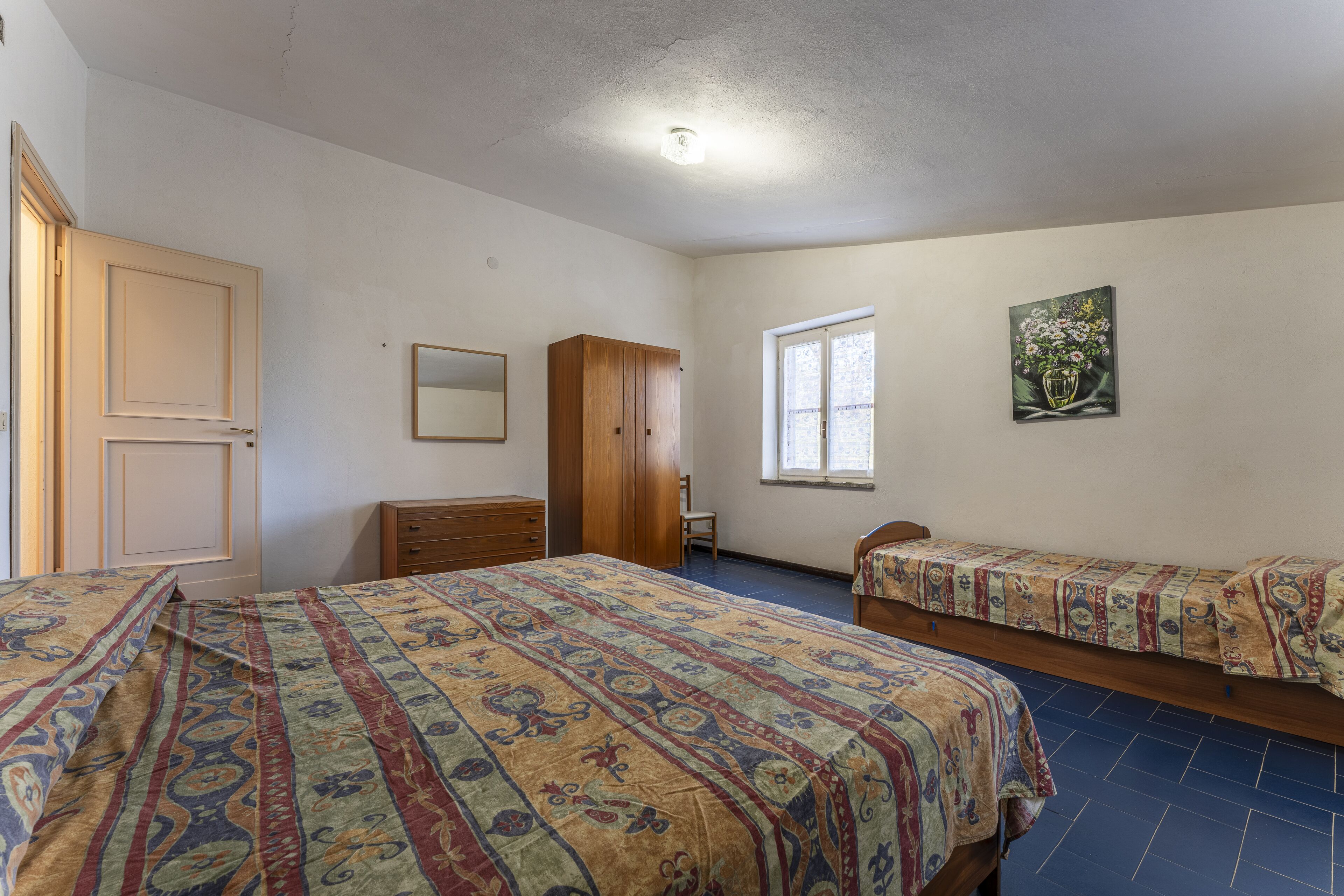 Quarto com duas camas, um guarda-roupa, penteadeira, janela e um quadro na parede.