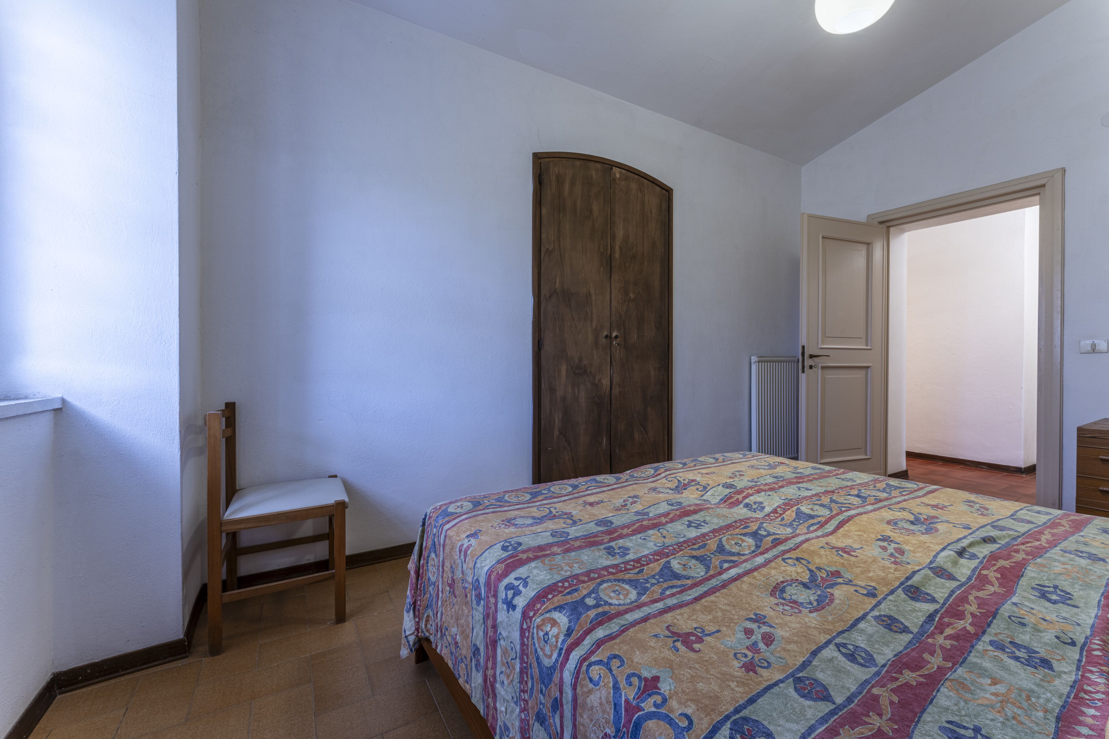Une chambre à coucher comprenant un lit, une chaise en bois, une armoire et une porte donnant sur une autre pièce.