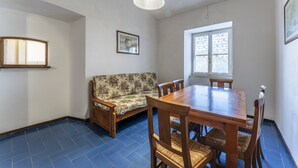 A room with a wooden dining table, chairs, a floral sofa, a window with patterned shutters, and a hanging light fixture. - Orbetello Giannella Apartment (Orbetello)