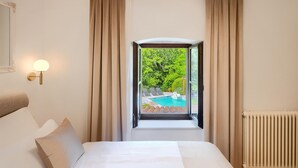 Une chambre dotée d’un grand lit, de rideaux beiges, d’une vue sur une piscine extérieure et d’un luminaire fixé au mur.