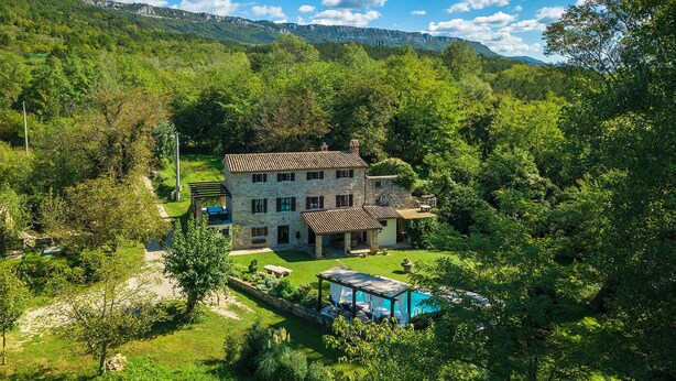 Une maison en pierre avec un toit de tuiles, entourée d’une végétation luxuriante et dotée d’une piscine.