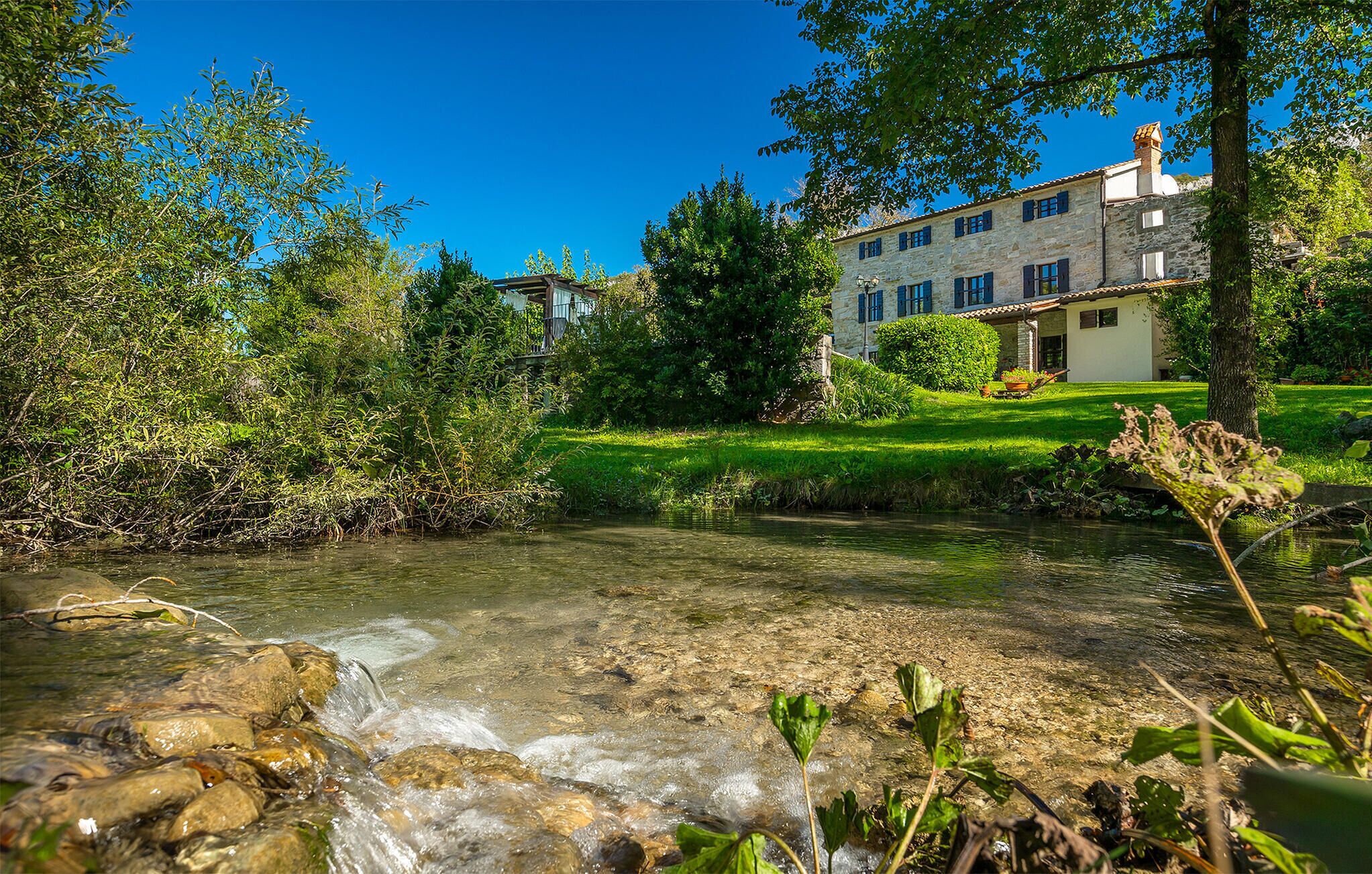 Un arroyo cristalino fluye por un exuberante jardín con un edificio de piedra al fondo.