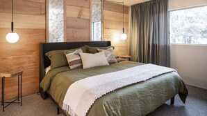 Een slaapkamer met een houten hoofdeinde, twee nachtkastjes, een bed met groen beddengoed, een witte plaid en een raam met gordijnen.