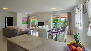 Un salon moderne avec un canapé, une table à manger pour quatre personnes et un coin cuisine agrémenté d’un panier de fruits.