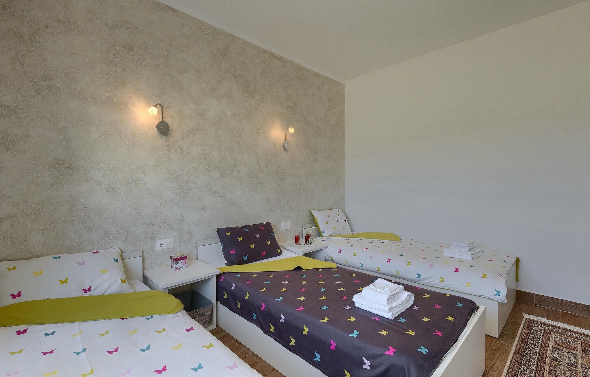 Ein Hotelzimmer mit zwei Einzelbetten, beide mit farbenfroher Schmetterlingsdecke, einem kleinen Nachttisch mit Lampe und einer schlichten Wand mit zwei Wandleuchten.