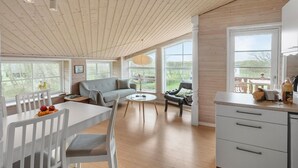 Amplia sala de estar con techo de madera, ventanas grandes, un sofá, una mesa de comedor con sillas y una cocina.