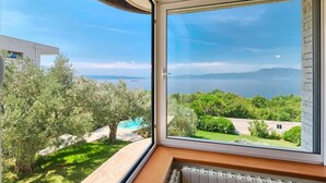 A room with a large window overlooking a pool and greenery, with a view of the sea and mountains in the distance.