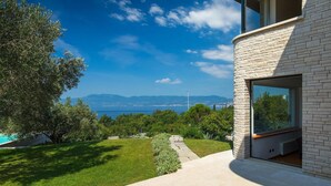A modern house with a stone facade, a large glass window, and a view of the sea and mountains.
