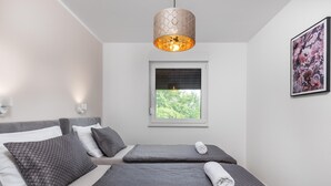 A modern bedroom with two beds, a window with blinds, a pendant light, and a framed floral artwork on the wall.