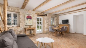 A cozy living room with a stone wall, wooden beams, a grey sofa, a dining table with chairs, and a TV on a wooden cabinet.