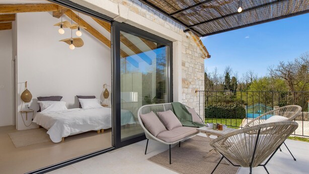 A modern bedroom with a glass door leading to a balcony, a wicker chair, and a small table with a book and glasses.