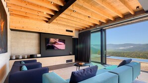 A modern living room with a large TV, comfortable seating, and a scenic view through large windows.