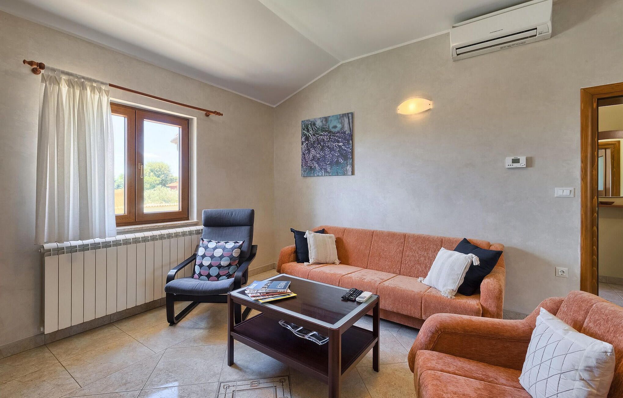A living room with a sofa, armchair, and coffee table, a radiator, an air conditioning unit, and a window with curtains.