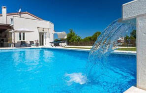Ein Swimmingpool mit glatter, blauer Wasseroberfläche, ein weißes Haus im Hintergrund und eine Springbrunnen-Anlage.