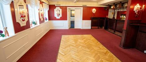 A hotel lobby with a red carpet, wooden herringbone floor, white wainscoting, chandeliers, and a bar area with a wine rack.