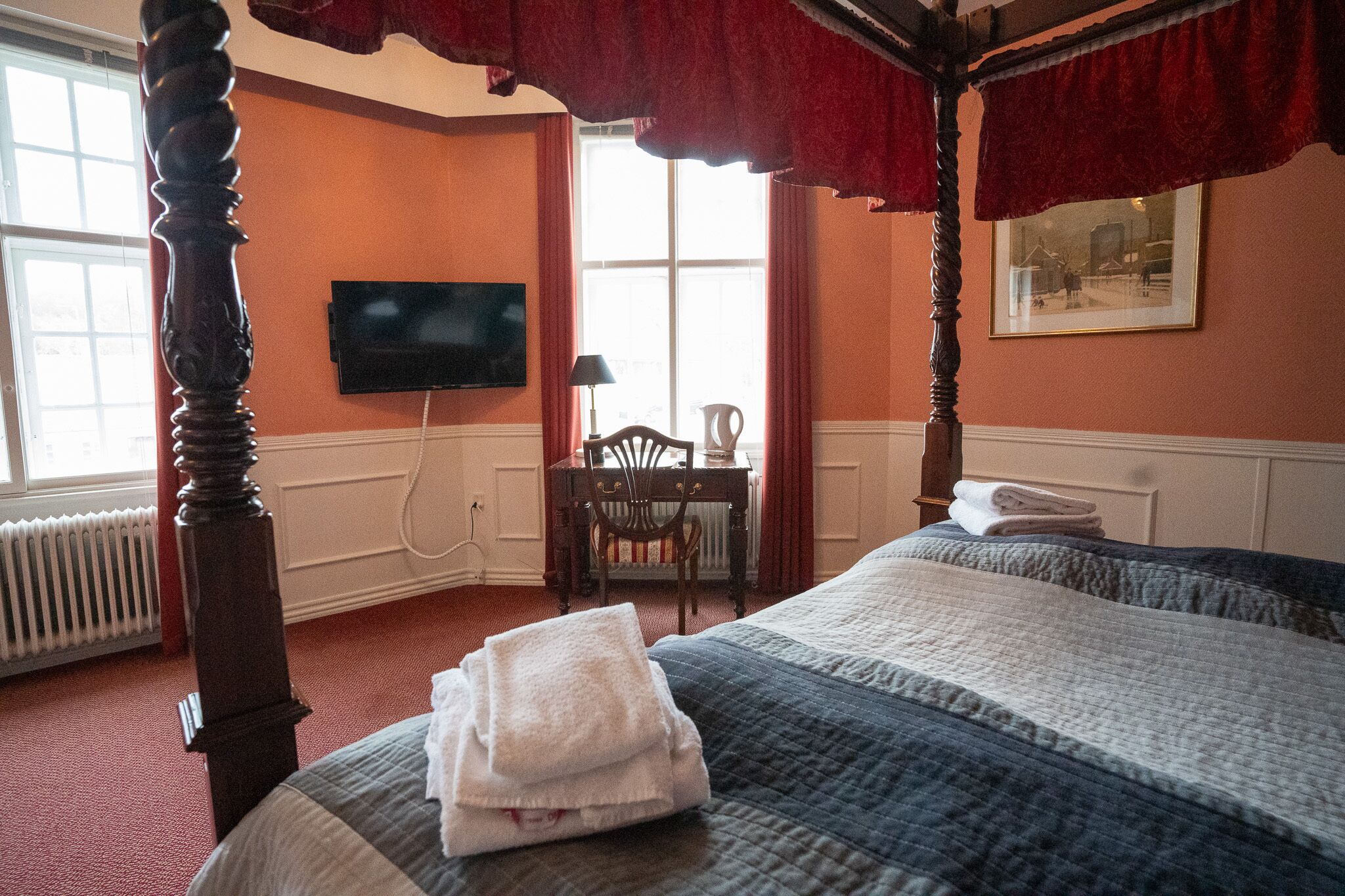 A four-poster bed with a folded towel, a television on the wall, a chair, and a window with curtains.