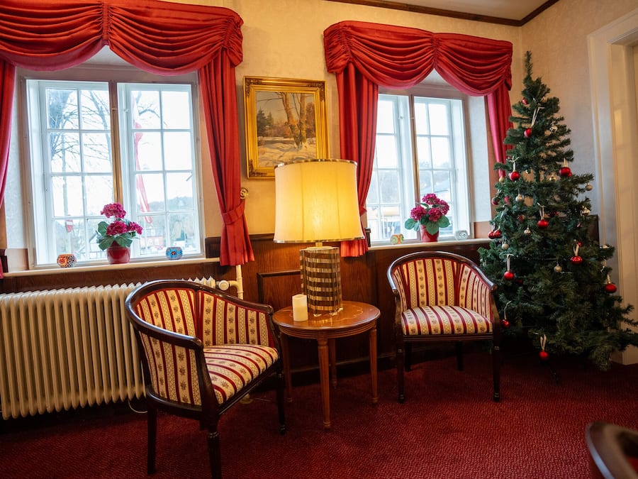 Um quarto aconchegante com duas poltronas estofadas, uma mesa com abajur, um radiador e um pinheiro de Natal.