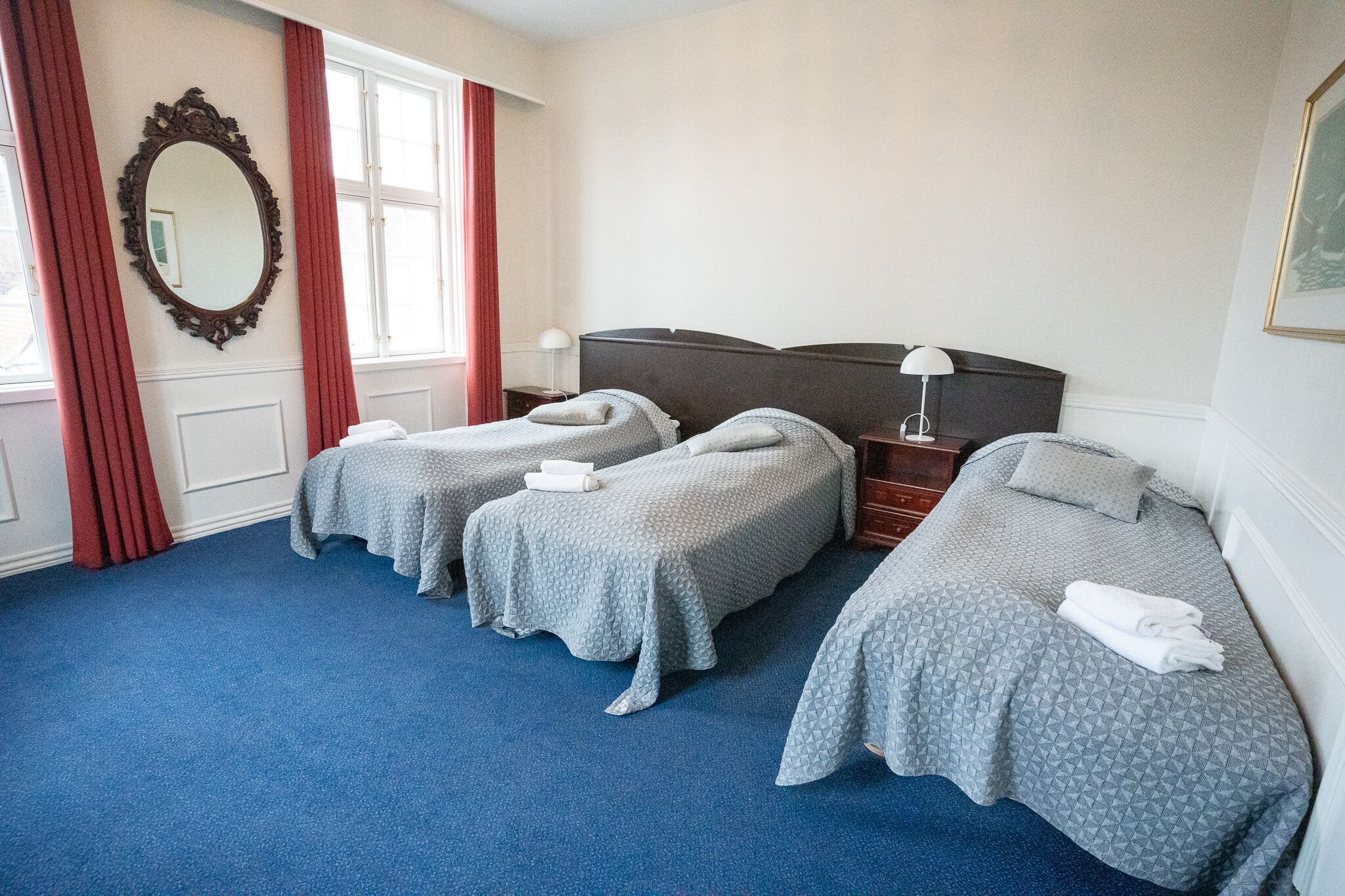 A hotel room with two single beds, a large mirror, a window with red curtains, and a blue carpet.
