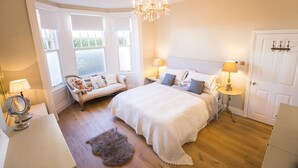 A bedroom with a large bed, two bedside tables with lamps, a bench with pillows, a chandelier, and a door.