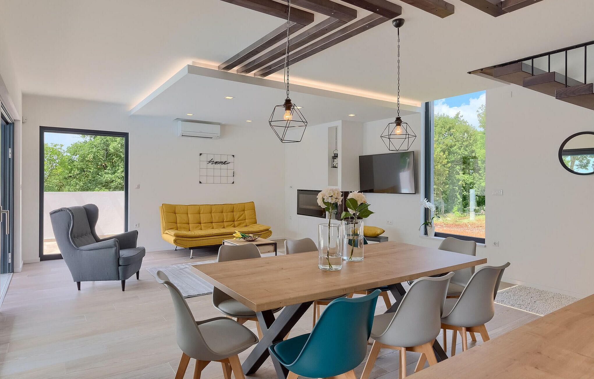 A modern living space with a wooden dining table, grey and blue chairs, a yellow sofa, and a TV mounted on the wall.