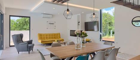 A modern living space with a wooden dining table, grey and blue chairs, a yellow sofa, and a TV mounted on the wall.