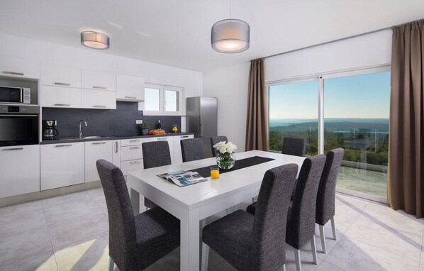 A modern kitchen with white cabinets, a dining area with a white table and grey chairs, and a large window offering a scenic view. - Nice home in Labin - Rabac with sauna (Labin - Rabac)
