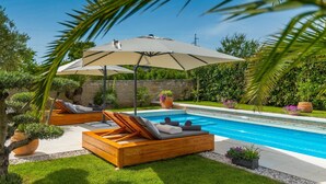 A poolside area with wooden loungers, a large umbrella, and potted plants.