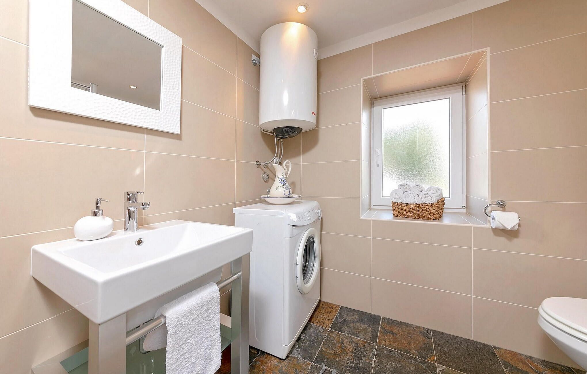 A modern bathroom with a sink, washing machine, and a window with a basket of towels.