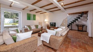 A spacious living room with a high ceiling, exposed wooden beams, a large window, a television, and a staircase leading to an upper floor.