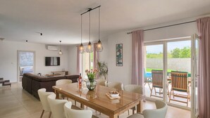 Ein modernes Wohnzimmer mit einer Couch, einem Esstisch mit Stühlen, einem an der Wand befestigten Fernseher und Blick ins Freie durch eine Glastür.