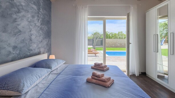 Ein Schlafzimmer mit Bett, Kissen und Blick auf ein Schwimmbecken durch eine Glastür.