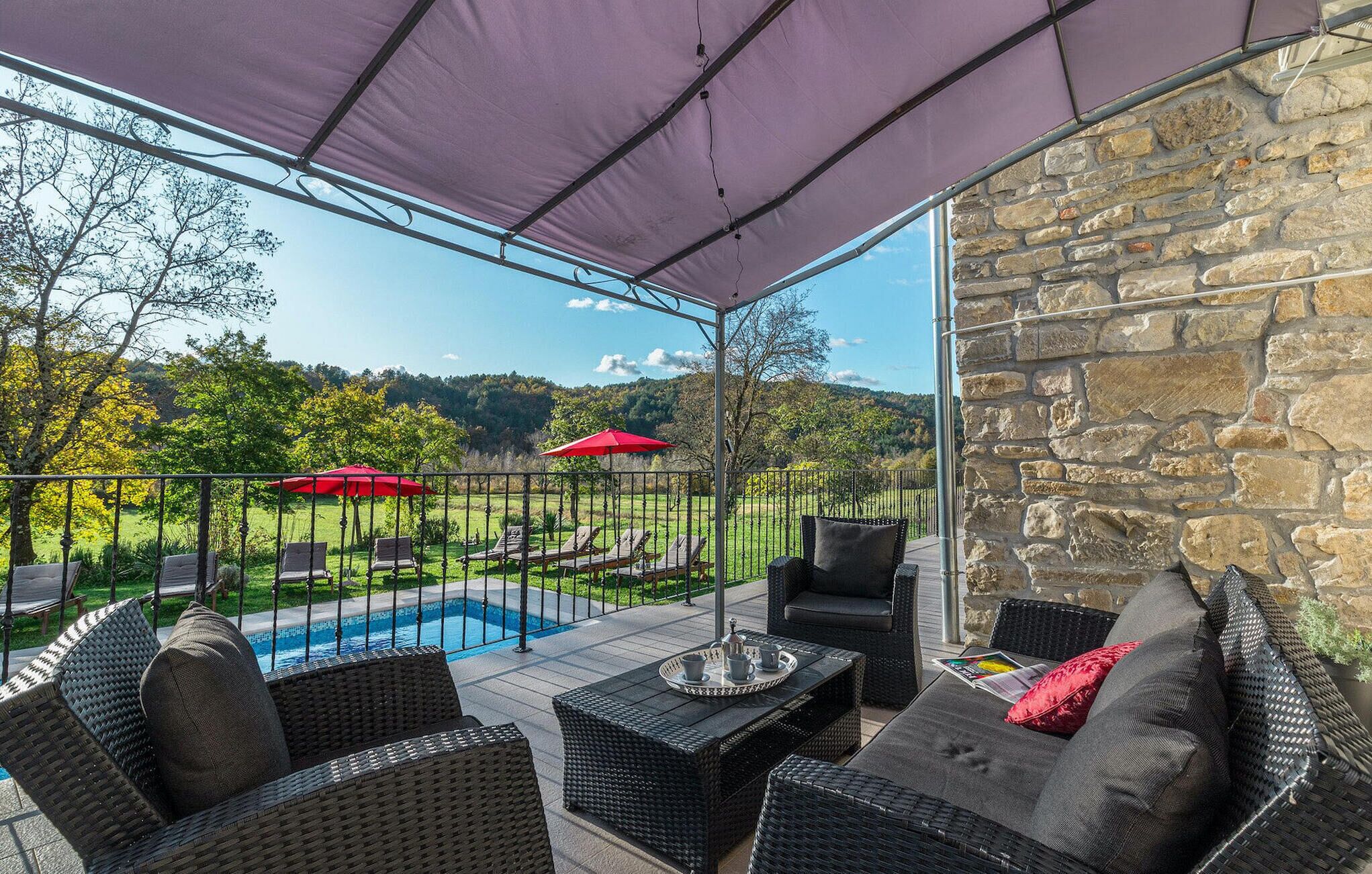 A covered patio with wicker furniture, a pool, and a stone wall.