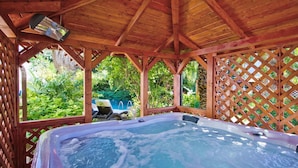 A hot tub under a wooden gazebo with a view of a swimming pool and lush greenery.
