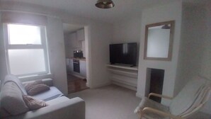A small, well-lit living room with a sofa, a TV, and a chair. There is a kitchen in the background.