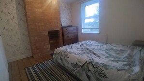 A bedroom with a brick fireplace, a bed with a floral bedspread, a dresser, and a window with blinds.