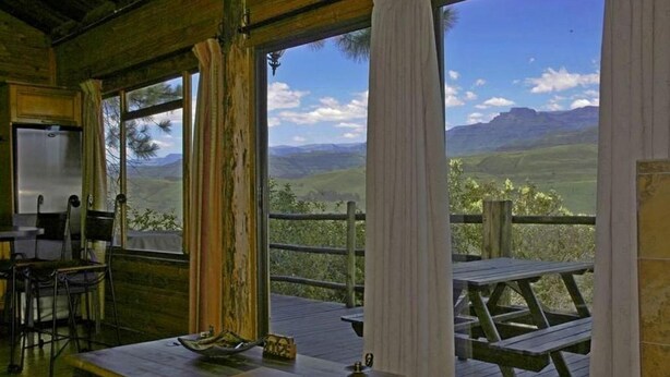 A rustic cabin with a wooden interior, a dining table, and a view of a mountainous landscape through large windows.