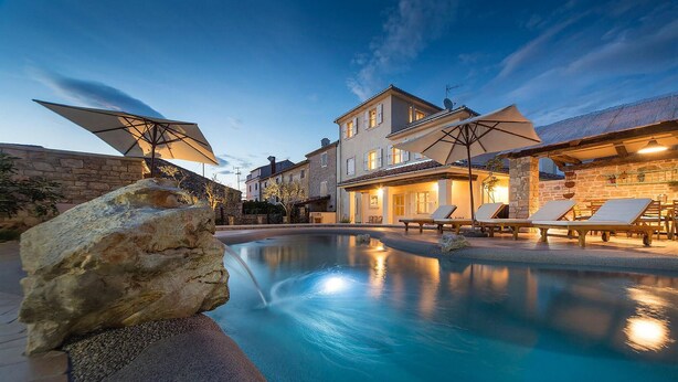 A swimming pool with lounge chairs and umbrellas, surrounded by stone walls and buildings.
